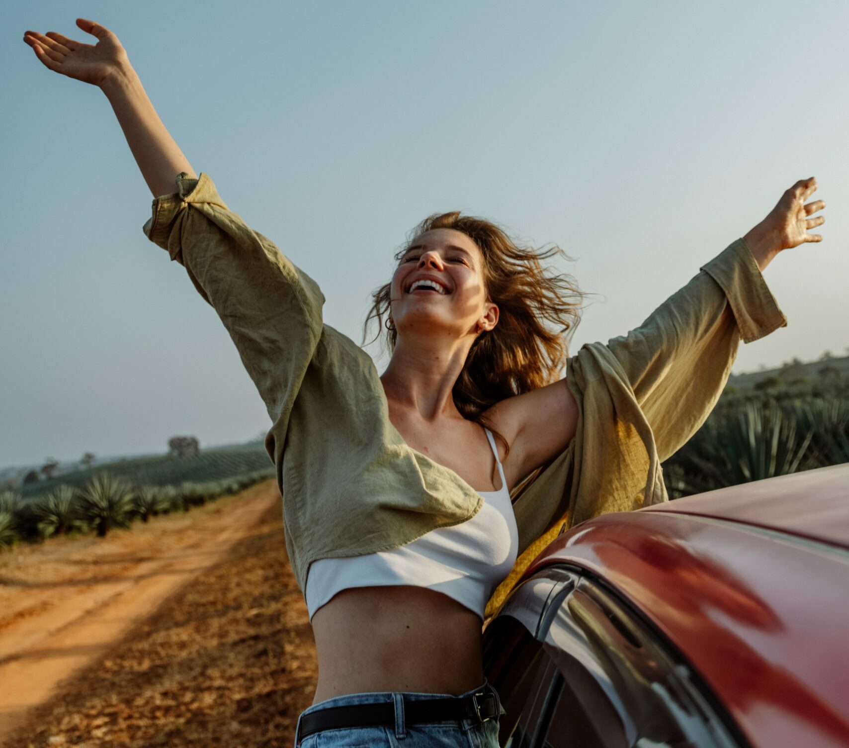 A woman with her arms raised to the sky, face looking up to the sky and smiling. Perched on the windowsill of a car. Wind blowing in her hair.