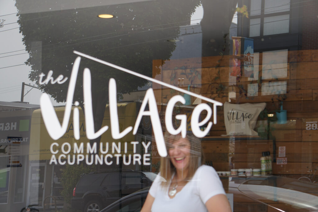 Sara Ward, standing behind a window, laughing . With the signage of The Village Acupuncture.