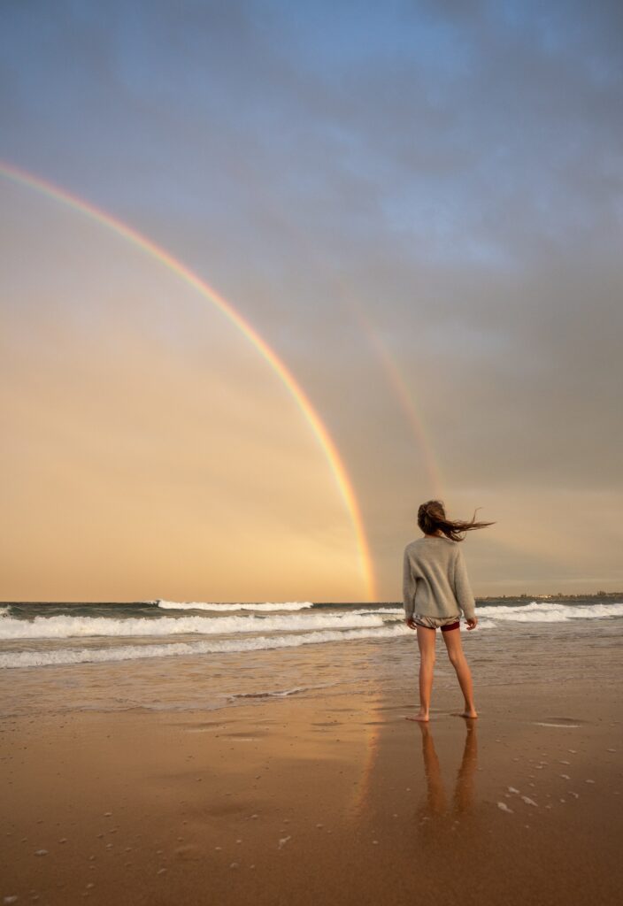 girl standing on a beach with a rainbow in the distance